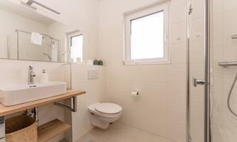 A modern bathroom with a shower, a sink on a wooden shelf, and a toilet area. Bright walls and a window create a friendly atmosphere.
