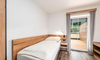A simple bedroom with a single bed and wooden details. Large windows offer a view outside and plenty of daylight.
