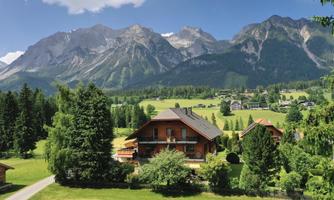 A picturesque landscape with a wooden house in the foreground and majestic mountains in the background. Lush green meadows and clusters of trees enhance the image.
