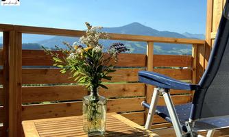 A cozy balcony with a flower vase on a wooden table. In the background, mountains and a clear sky can be seen.