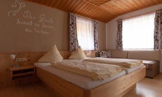 A cozy bedroom with a large bed and wooden decor. The walls are bright, and large windows provide plenty of light.