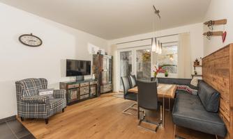 A modern living room with wooden flooring and large windows. There is a cozy seating area, a dining area, and a stylish decor.