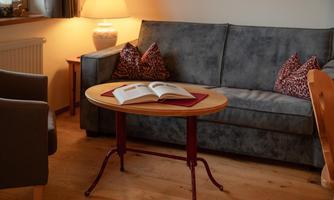 A cozy living room with a gray sofa and a round table. On the table lies an open book and a decorative lamp stands in the background.