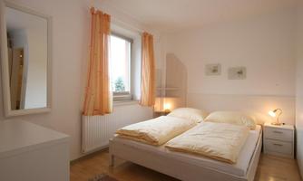 A bright bedroom with a double bed and large windows. The walls are painted white, and there are some decorative elements.