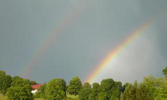 A beautiful rainbow arches over a green landscape with trees. In the background, a house can be seen under a cloudy sky.