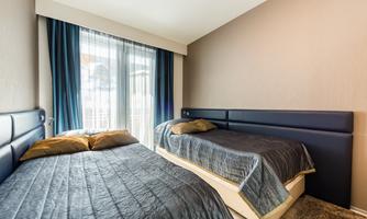 A modern bedroom with two single beds and a large window. The walls are in neutral colors and the bedding is dark.