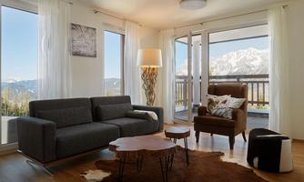 A modern living room with large windows and a view of the mountains. There is a sofa, an armchair, and a unique wooden coffee table.