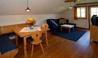 A cozy living kitchen with a wooden table, chairs, and a blue sofa. A bouquet of flowers is on the table, and the windows provide plenty of natural light.