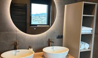 A modern bathroom with two sinks and a round mirror. Nearby are fresh towels and a window offers a view of the surroundings.