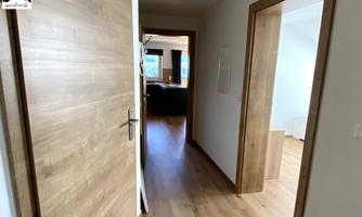 A bright corridor with wooden flooring and open doors to adjacent rooms. The walls are painted in neutral colors.