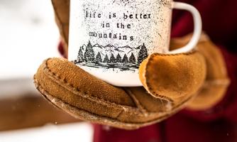 A person is holding a cup in their hands, which says: "Life is better in the mountains." The cup is white with a forest motif, and in the background, snow-covered landscapes are visible.