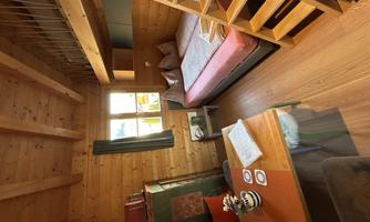 A cozy room made of wood with a double bed and a table. Lots of natural light comes in through the windows.