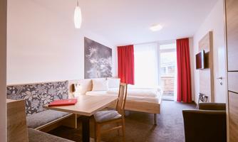 A cozy hotel room with a large bed and a seating area. The bright colors and the windows with red curtains create an inviting atmosphere.