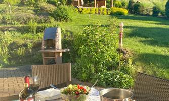 A table outdoors with grilled meat, salad, and red wine. In the background, there is a beautiful meadow and a garden gazebo.