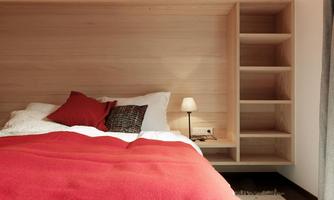 A modern bedroom with a wooden panel in the background. The bed is decorated with red pillows and a blanket, next to it stands a lamp and a shelf.