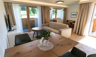 A modern living room with a comfortable couch and a large dining table.  
Much daylight falls through the windows and you can see a beautiful view of nature.