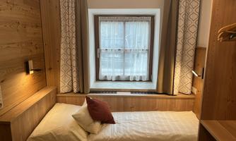 A cozy bedroom with wooden walls and a large window. A simple bed with pillows and a decorative cushion is available.