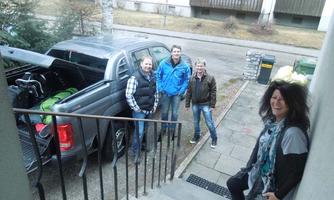 A group of four people stands in front of a vehicle loaded with luggage. In the background, there is a residential building and a quiet street.