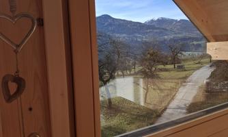 A view from a wooden window onto a picturesque landscape with mountains in the background. In the foreground, trees and a path can be seen.