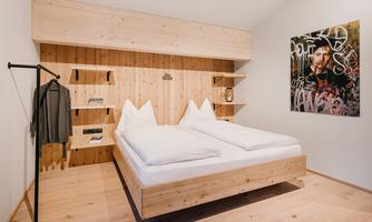 A modern bedroom with a double bed and wooden accents. A graffiti-based artwork hangs on the wall, and there is a clothes rack as well as shelves.