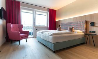 A modern hotel room with a large bed and a pink armchair. The windows provide plenty of daylight and overlook a balcony.