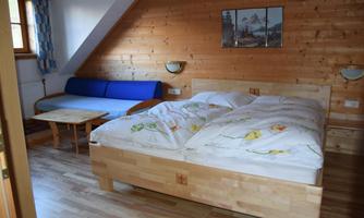 A simple bedroom with a large double bed and a blue sofa. Additionally, there is a small table and wood paneling on the walls.