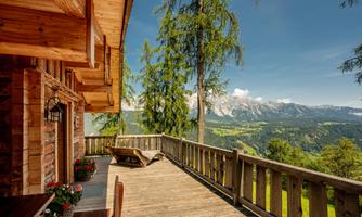 A cozy wooden cabin with a view of an impressive mountain landscape. The terrace is surrounded by trees and flowers, perfect for relaxing moments in nature.