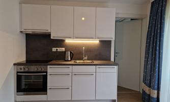 A modern kitchen with white furniture and a tasteful countertop. It is well-lit and includes a stove, a sink, and a kettle.
