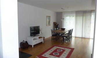 A modern living room with a television, a large table, and chairs. The room has a wooden floor and a decorative carpet pattern.