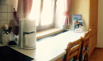 A bright kitchen with a white kettle and a stack of wooden chairs. On the countertop is a magazine and the windows are adorned with curtains.