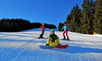 Three people on a ski slope in sunny weather. A snowboarder is sitting on the slope while the other skiers are standing nearby.