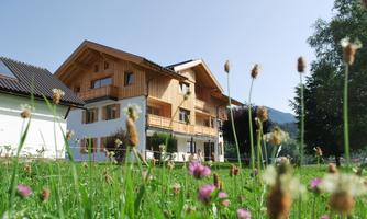 A modern wooden house surrounded by green meadows and colorful flowers. The clear sky and nature give the scene a calming atmosphere.