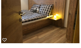 A cozy bedroom with a bed and a bedside table. The walls and furniture are made of light wood, and a soft light creates a warm atmosphere.