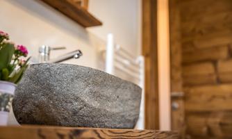 An elegant natural stone sink stands on a wooden table. In the background, a plant and a towel holder can be seen.