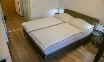 A comfortable double bed with white bedding in a bright room. The floor is made of light wood, and there is a simple bedside lamp.
