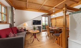 A cozy living room with a wooden ceiling and large windows. There is a seating area with a sofa and a dining table for four people.