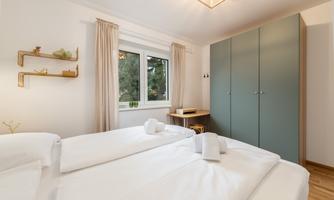 A cozy bedroom with two single beds and white bed covers. On the wall, there is a window, curtains, and a green wardrobe.