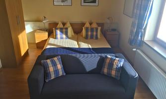 A cozy bedroom with a double bed and two pillows. In the foreground, there is a sofa with checked cushions.