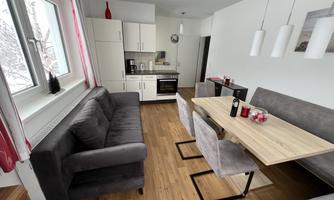 A modern living room with a gray couch and a wooden dining table. The space is bright and cheerful, with an open kitchen in the background.