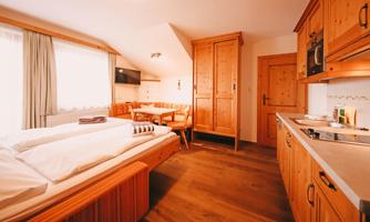 A cozy room with wooden paneling, a double bed, and a small kitchenette. There is a dining area with a table and chairs, as well as a window with plenty of natural light.