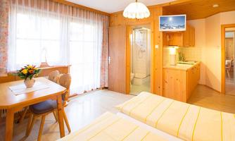 A cozy room with two beds, a table, and a modern kitchen. Large windows provide bright light and an inviting atmosphere.