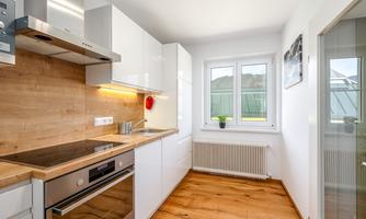 A modern kitchen with white high-gloss cabinets and a wood countertop. The large windows provide plenty of daylight.