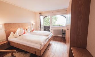 A bright and cozy hotel room with two single beds and wooden details. The room has a window overlooking the outside and is simply furnished.