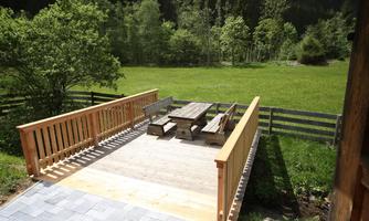 A spacious wooden terrace with a large table and benches. In the background, you can see a green meadow and trees.