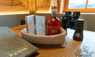 A cozy table with a menu, a bottle of ZIRAV, and a pair of binoculars. In the background, wooden walls and windows are visible, creating a warm atmosphere.