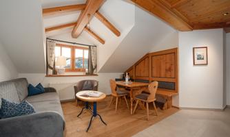 A cozy attic room with wooden beams and a large window. The decor includes a sitting area with a sofa and a dining area with chairs.
