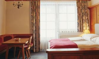 A cozy room with a double bed and a table with chairs. Large windows let in plenty of daylight and give the space a bright, friendly atmosphere.