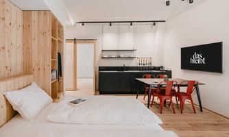 A modern room with wooden paneling and a cozy bed. The open kitchen and dining area with red chairs create an inviting atmosphere.