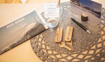 An invitation with a mountain motif, along with keys in a container and a handwritten note. A pen and a postcard are also on the table.
