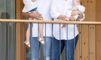 A happy family stands on the balcony of a wooden house. The parents are holding two small children while smiling.